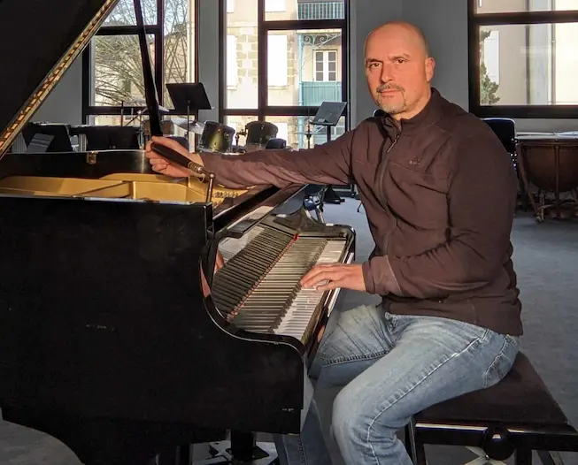 Michel BOURY , accordeur de piano professionnel assis à un piano à queue dans une pièce bien éclairée avec de grandes fenêtres en arrière-plan, accordant l'instrument avec concentration et précision.